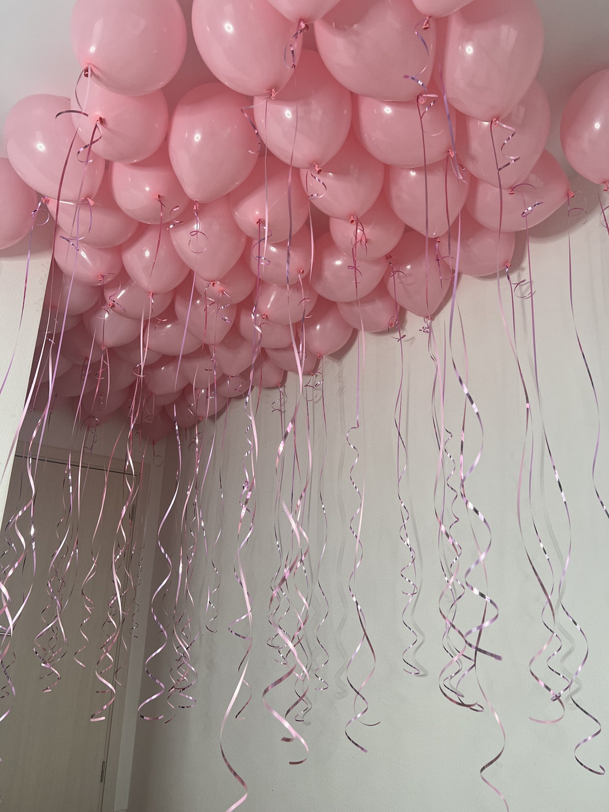 Baby pink helium balloons covering a white ceiling with long pink ribbons and curled ribbon details hanging below.