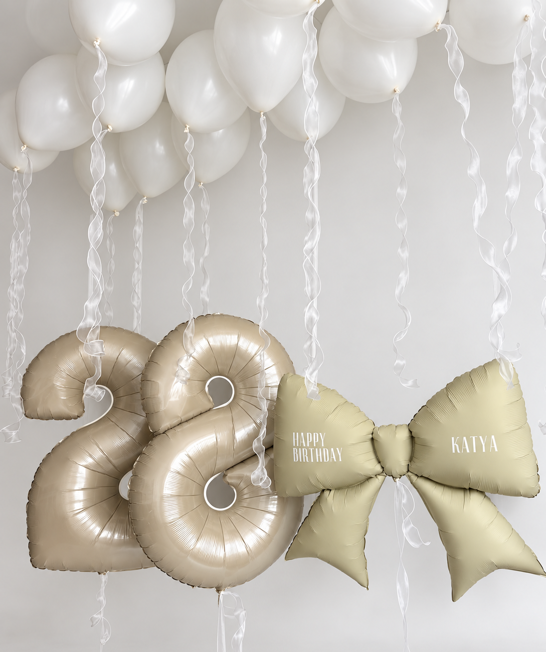 Two champagne number balloons beside a pale gold bow with personalized birthday text, beneath white ceiling balloons with flowing wavy ribbons.