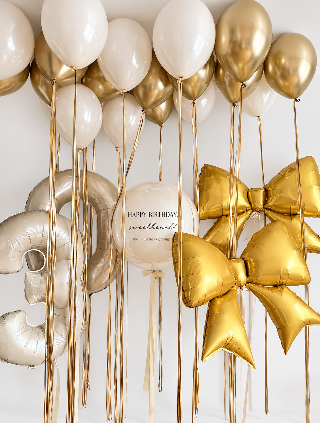 Cream and gold ceiling balloons with long gold ribbons above champagne number 30 balloons, a personalized cream bubble and two gold bow balloons.