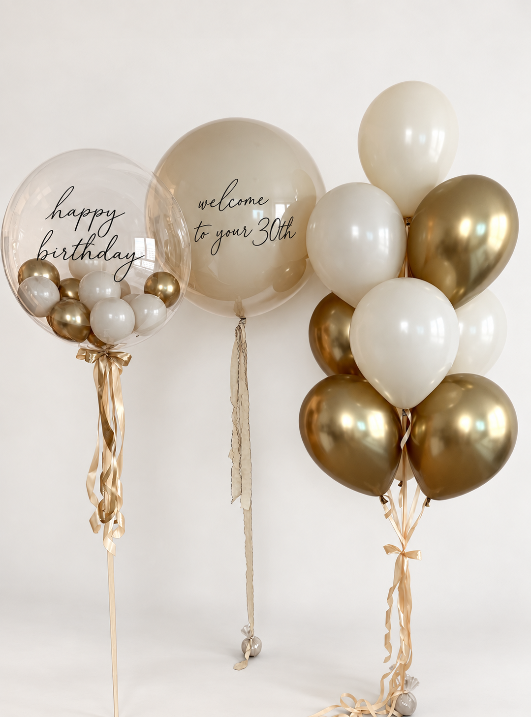 Two personalized bubble balloons with black birthday lettering, cream and gold balloon filling, and a bunch of cream and gold latex balloons with matching ribbons.