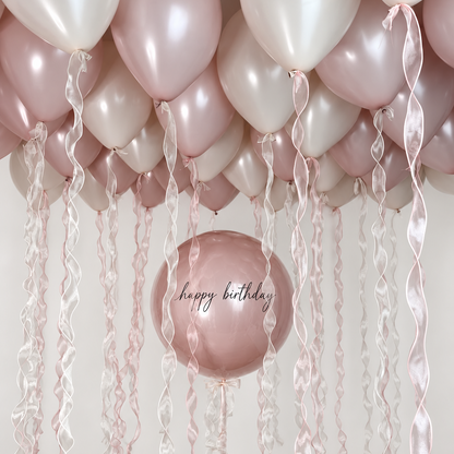 Personalized dusty rose bubble balloon with black happy birthday lettering beneath dusty rose and cream ceiling balloons with pink and white wavy ribbons.