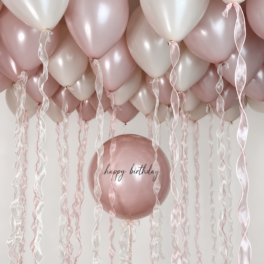 Personalized dusty rose bubble balloon with black happy birthday lettering beneath dusty rose and cream ceiling balloons with pink and white wavy ribbons.