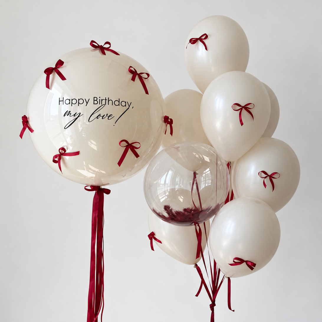 Large personalized cream bubble decorated with burgundy bows beside cream latex balloons with matching bows and a small clear bubble filled with burgundy feathers.