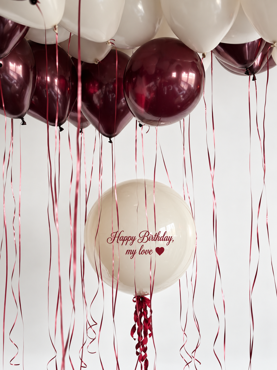 Personalized cream bubble balloon with burgundy birthday lettering beneath burgundy and cream ceiling balloons with long burgundy ribbons.