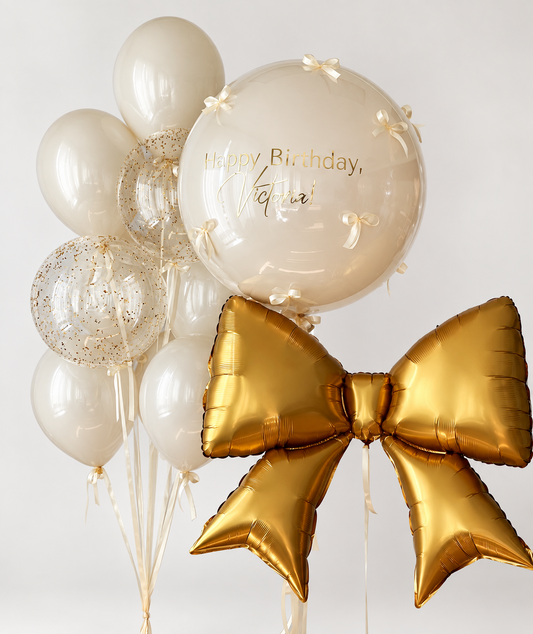 Large personalized cream bubble with small cream bows beside a gold foil bow and a bouquet of glossy cream balloons with two clear gold confetti bubbles.