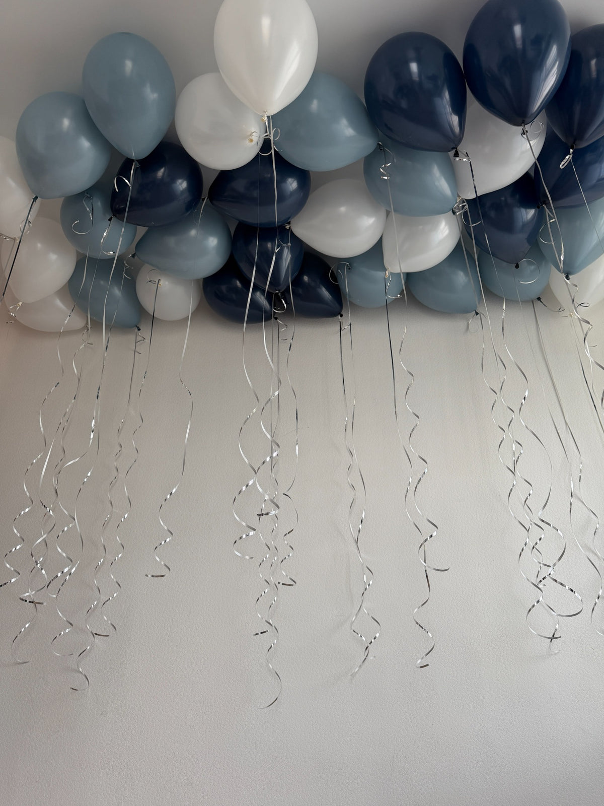 Navy, dusty blue and white helium balloons floating against a white ceiling with long silver ribbons and curled ribbon details.