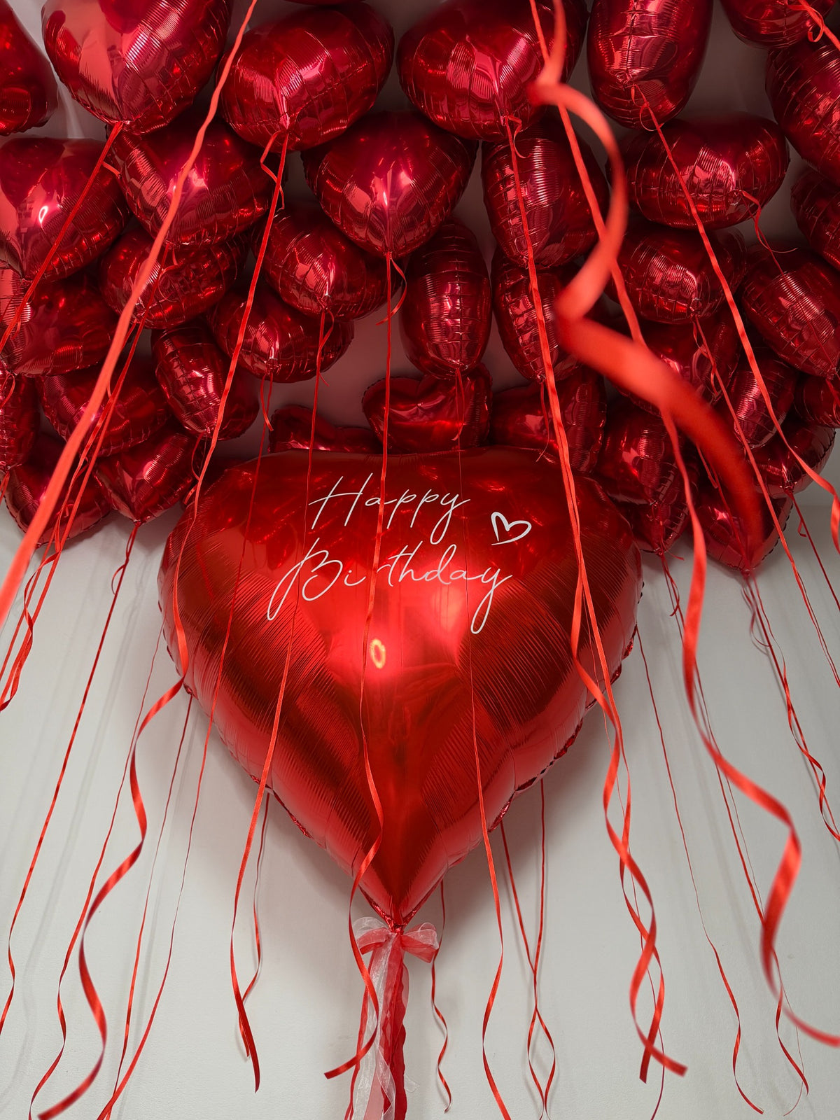 Red heart ceiling balloons with long red ribbons and a large personalized red heart balloon with white Happy Birthday lettering.