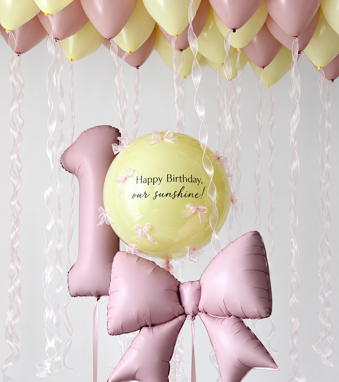 Dusty pink number one and bow balloons beside a personalized yellow bubble with small pink bows, beneath yellow and dusty pink ceiling balloons with wavy ribbons.